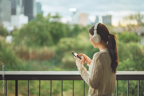 Young woman wearing headphones using smartphone on outdoor balcony, relaxing with music while enjoying nature view, modern lifestyle, technology and wellness concept