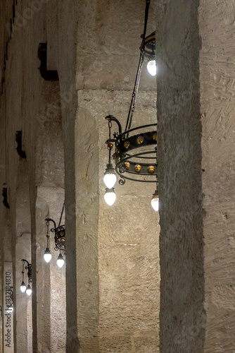 warm, atmospheric interior view of the Cathedral of Syracuse (Cattedrale di Siracusa) showing massive ancient stone columns illuminated by decorative hanging lamps. The image highlights the textured