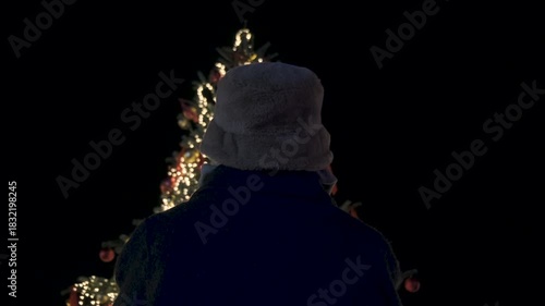Christmas ornaments on christmas tree in a christmas market. Alone unrecognizable woman looking to the tree. Celebration for new year. Lights and some bright decorations looking shiny. 