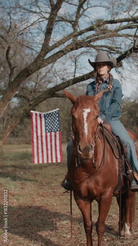Cowgirl Woman With Braid and Scarf Wearing Cowboy Hat and Denim Jacket Sitting On Brown Horse With American Flag and Tree in Field in the Background