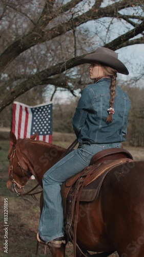 Cowgirl Woman With Braid and Scarf Wearing Cowboy Hat and Denim Jacket Sitting On Brown Horse With American Flag and Tree in Field in the Background