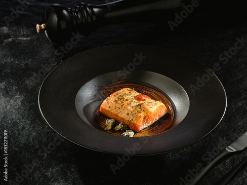A perfectly cooked salmon fillet rests in a stylish black bowl, adorned with herbs and served with artistic plating. The dish is presented in a setting ready for a delightful Dinner