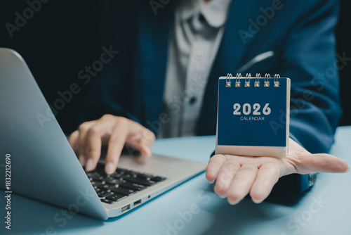 Small desktop calendar 2026 held in hand with person typing on laptop in background, business planning and organization concept with blue and gray tones