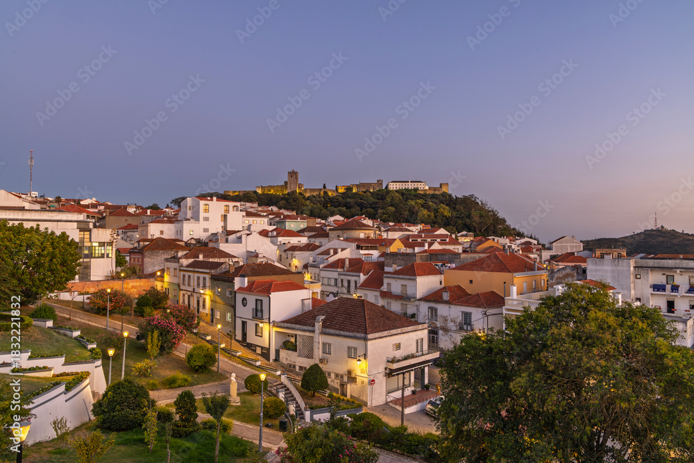 Obraz premium Palmela, Portugal, at dusk. The town lights are on, and the historic castle on the hill is illuminated against the soft twilight sky.