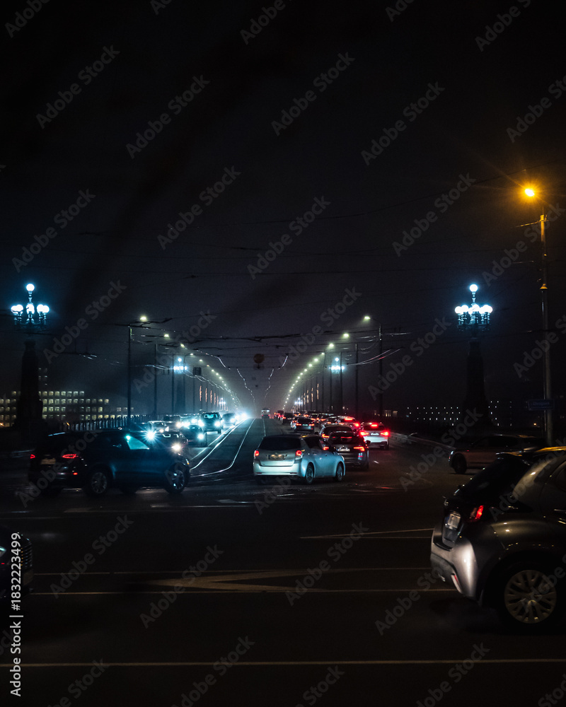 Fototapeta premium Nighttime city bridge with trams and cars moving through fog, illuminated by bright streetlights and overhead wires. The scene captures a moody urban atmosphere with soft light reflections.