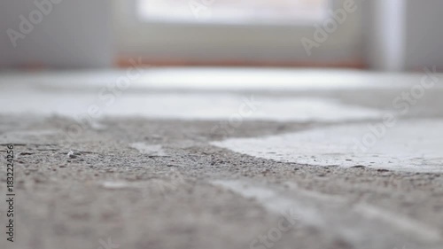Close-up of a dusty concrete floor being swept with a sweeping broom in an empty indoor space. Sunlight softly highlights the dust particles. Blurry background with a window