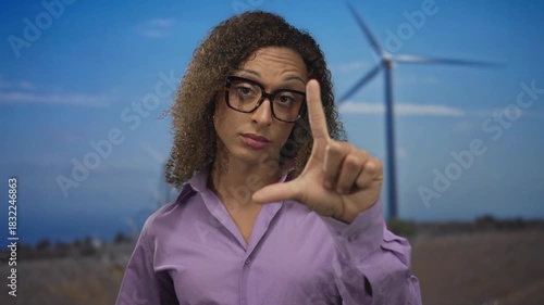 Woman points index finger and thumb in l shape at windmill in forest; mockery sarcasm critique disdain.