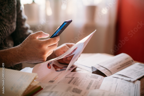 A man pays utility bills online using a QR code in an app using a smartphone in the kitchen. Close-up of hands, phone, and bills.