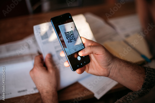 A man pays utility bills online using a QR code in an app using a smartphone in the kitchen. Close-up of hands, phone, and bills.