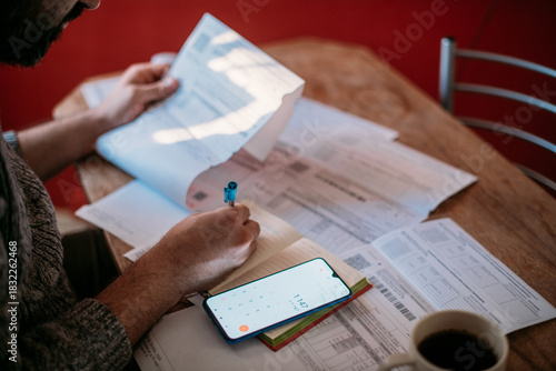 A man pays utility bills online using a QR code in an app using a smartphone in the kitchen. Close-up of hands, phone, and bills.