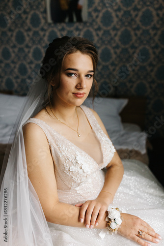 Portrait of a bride sitting indoors in her wedding dress. She looks at the camera with a calm, confident expression. Soft light highlights the lace details, veil, and floral bracelet, creating 