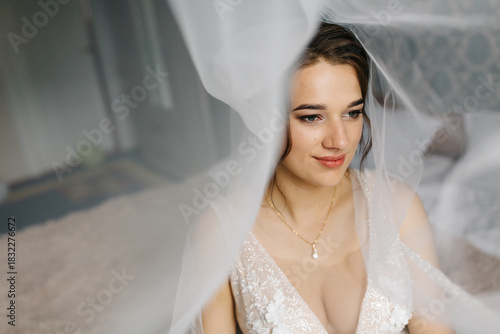 Bride smiling softly as her veil flows around her, creating a dreamy and romantic atmosphere. The close-up portrait highlights her makeup, lace wedding dress, and elegant jewelry in gentle natural 