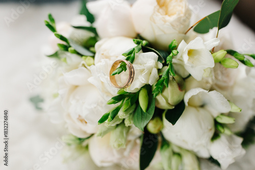 Close-up of wedding rings placed on a bouquet of white flowers. Soft natural light highlights the delicate petals, greenery, and golden bands, creating a romantic and elegant wedding detail shot.