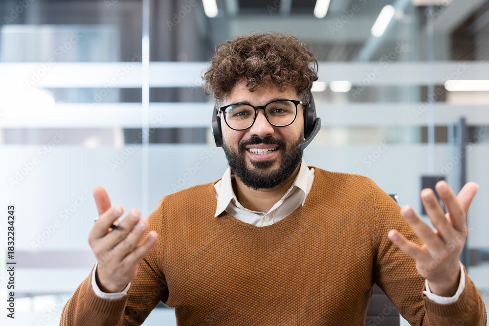 Obraz premium Smiling bearded man working as customer service in an office, wearing a headset and glasses, actively communicating and providing support during a video call