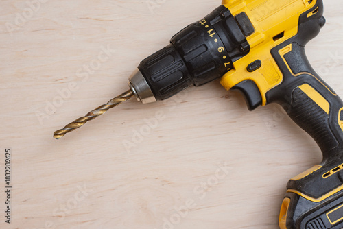 Yellow cordless drill with metal drill bit lying on light wooden background with empty copy space for text.