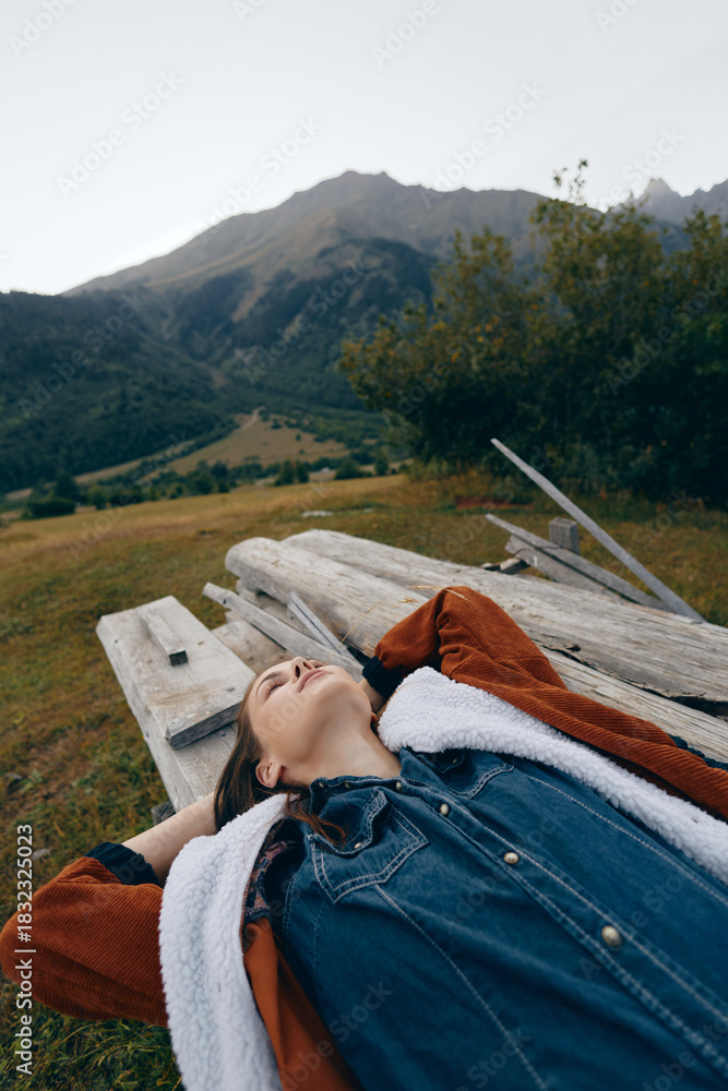 Fototapeta premium Woman relaxed on wooden bench outdoors in nature, portrait of casual traveler lying back in denim and shearling jacket on meadow with mountain backdrop and peaceful expression