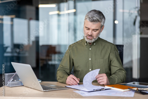Mature professional businessman is reviewing contracts and legal papers while working at his desk in a modern office, signing important documents with a pen