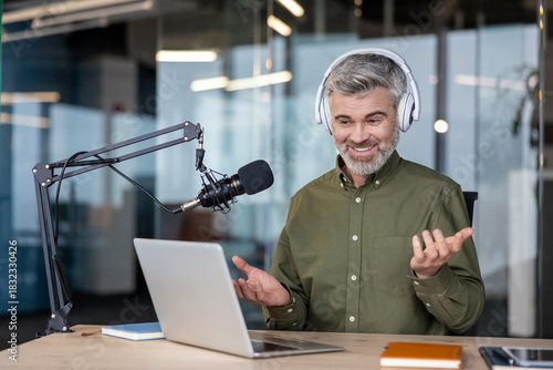 Mature man wearing headphones and using a studio microphone with a laptop, broadcasting or recording an online podcast from a professional office environment, actively gesturing while speaking