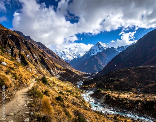 Scenic mountain valley landscape with a winding trail, river, and snow-capped peaks under a blue sky with clouds