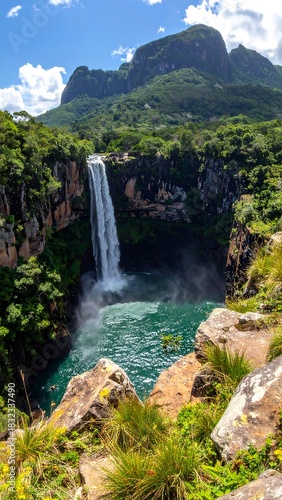 Waterfall plunges into turquoise pool surrounded by verdant cliffs and a distant mountain range under a partly cloudy sky