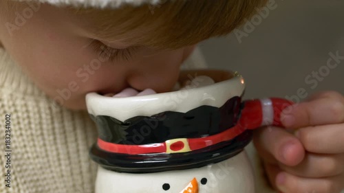 Cozy moment with boy enjoying warm cocoa during winter season. Boy leaning close to steaming cocoa in snowman mug, capturing charming Christmas holiday atmosphere with gentle lights, sweet flavors.