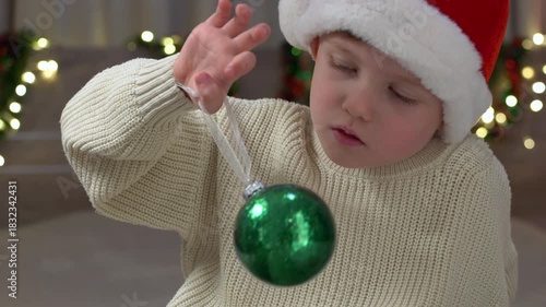 Little boy in Santa hat holding shiny green ornament, interacting with playful curiosity in warm holiday setting, surrounded by festive lights, capturing magical childhood atmosphere with soft colors.