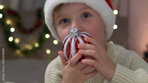 Young boy with Christmas decoration and warm glow, young boy wearing santa hat with sparkling ornament and cozy ambiance, child dressed as santa with bright bauble amid soft lights and holiday warmth