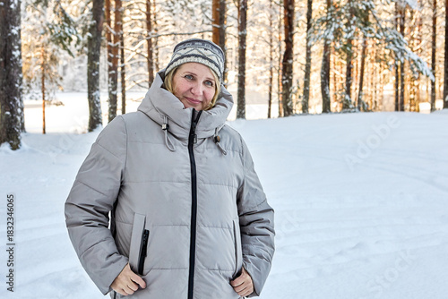Warmly dressed mature woman stands in snowy forest while her companion takes photo.