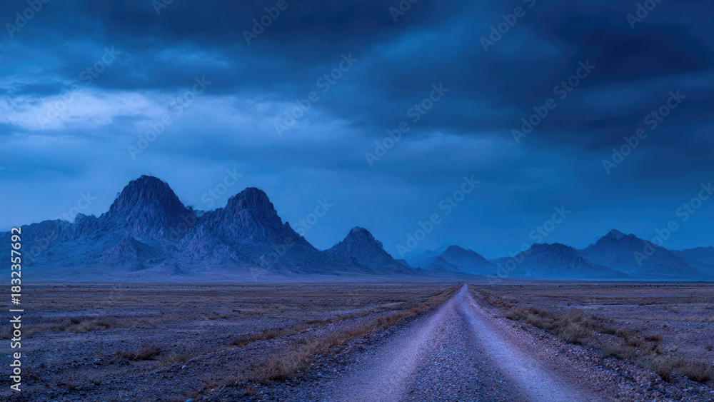 Fototapeta premium A long, solitary gravel road extends through a vast, arid landscape towards majestic blue mountains under a dramatic, cloudy twilight.