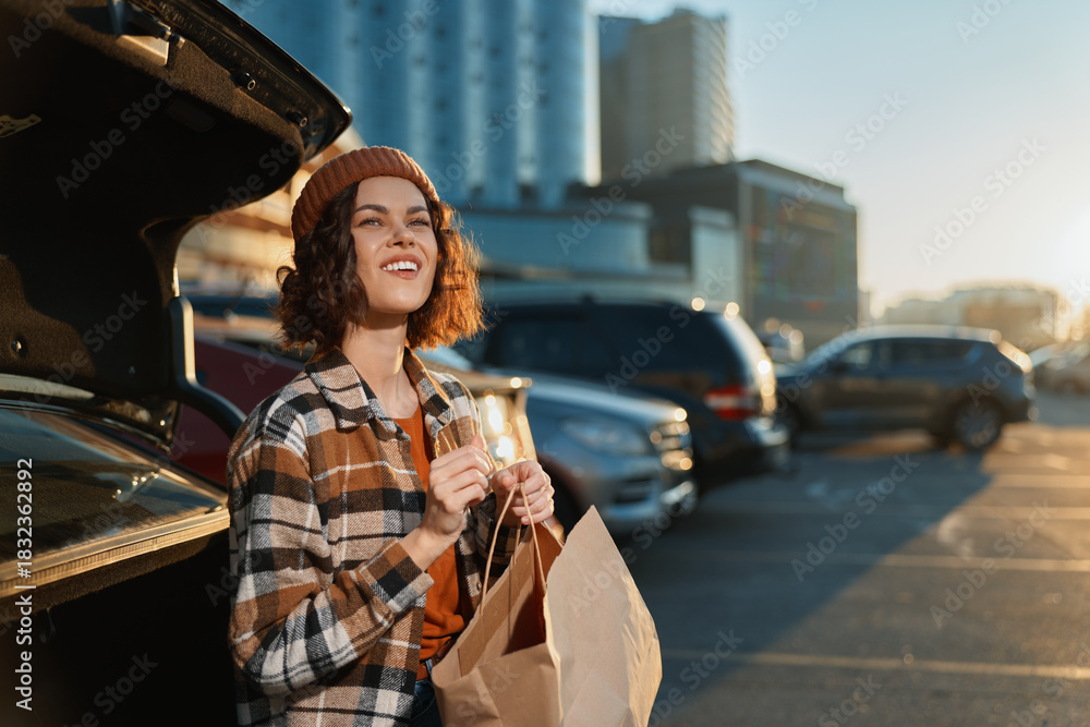 Fototapeta premium Woman by car in parking lot holding a paper shopping bag, smile and sunlight at golden hour glow. Candid lifestyle portrait with authenticity, mindful living and emotional storytelling vibe.
