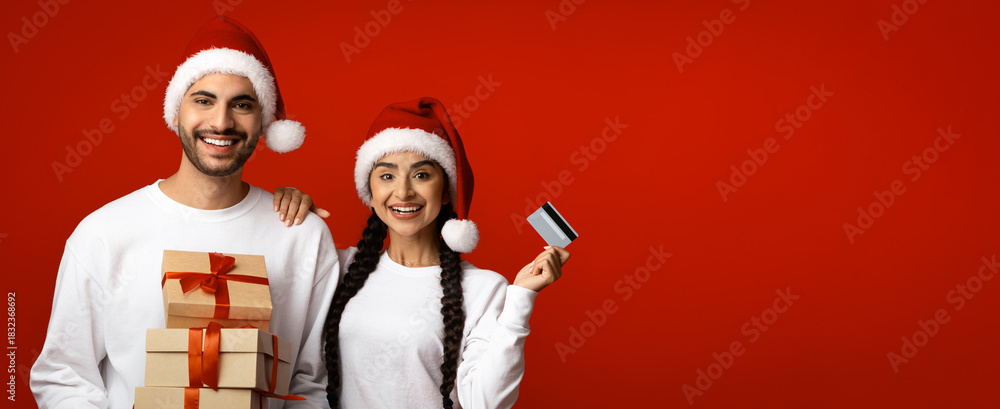 Naklejka premium Two people are smiling joyfully while wearing Christmas hats. One holds a stack of wrapped presents, while the other displays a credit card, showcasing holiday spirit and excitement.