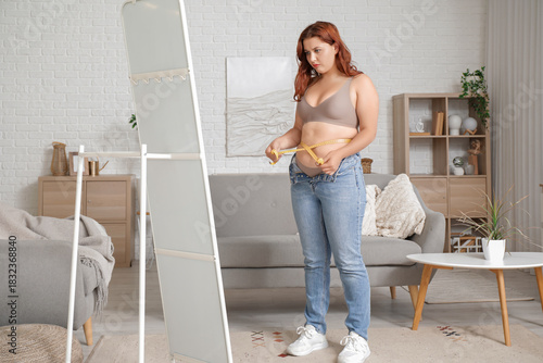 Overweight young woman in tight jeans with measuring tape near mirror at home...