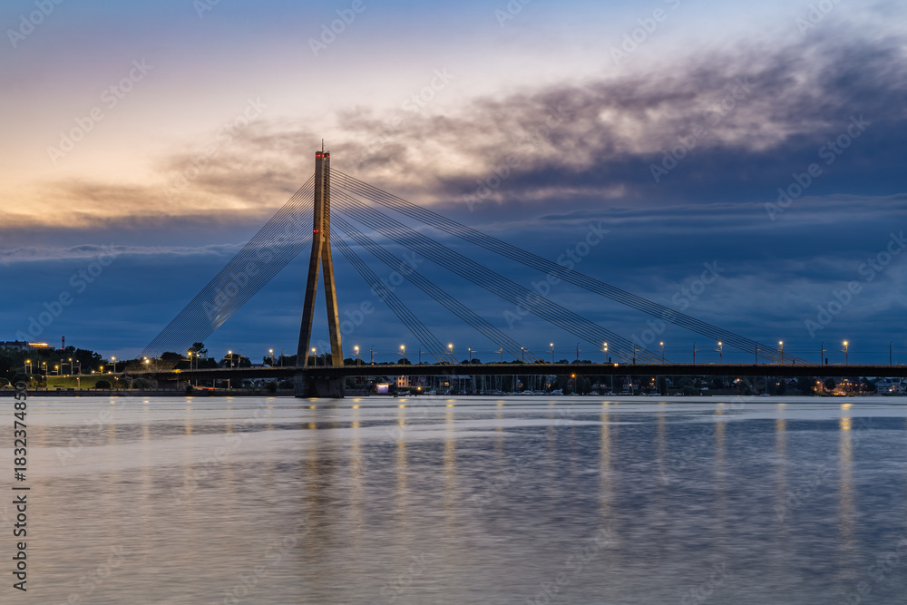 Obraz premium The illuminated Vansu Bridge, a cable-stayed bridge, spans the Daugava River at twilight, with its reflection visible on the calm water and a dramatic cloudy sky overhead