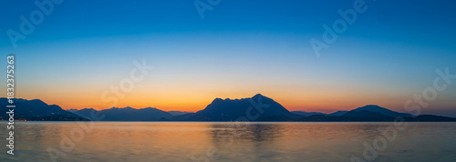 Panoramic view of Lake Maggiore and the silhouette of the surrounding Alps and mountains (likely Mottarone), reflecting the vibrant orange and blue colors of dawn or dusk from Stresa