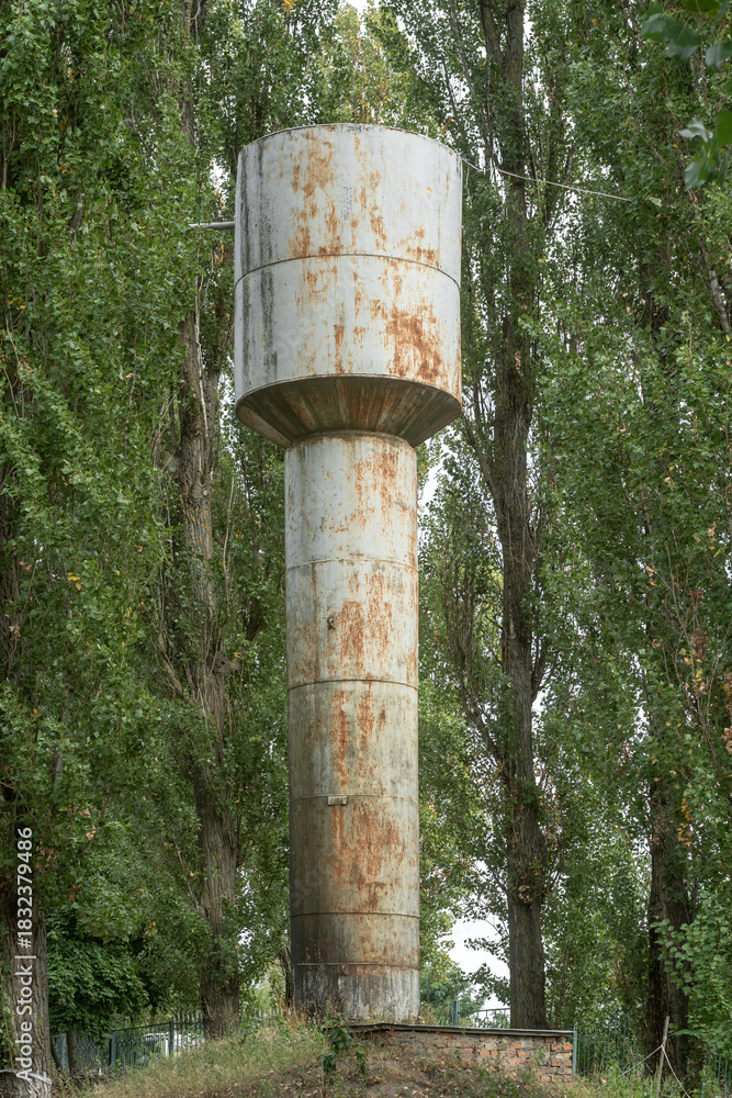 Fototapeta premium Rusted cylindrical water tower among tall poplar trees on a small hill.