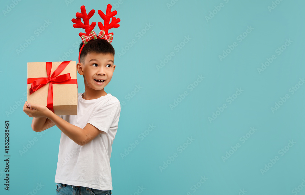 Fototapeta premium A cheerful child wearing festive reindeer antlers is carrying a wrapped gift box. The bright blue background creates a joyful holiday atmosphere.