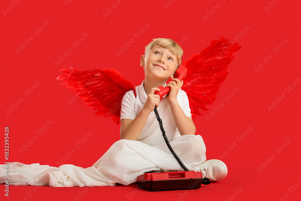 Obraz premium Cute little happy boy dressed as cupid with retro phone sitting on red background. Valentine's Day celebration