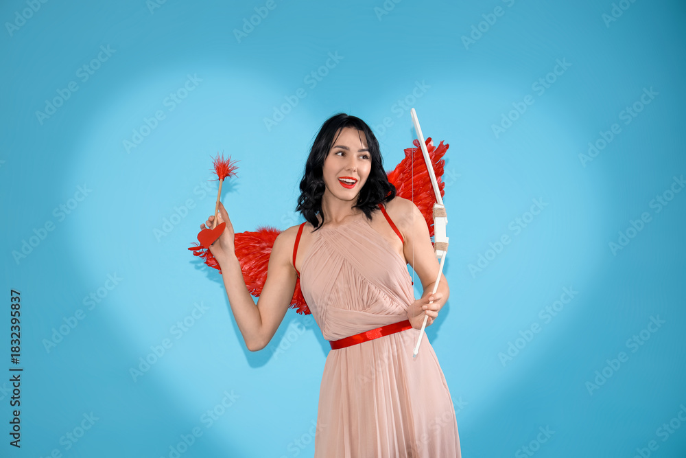 Fototapeta premium Beautiful young happy woman dressed as cupid with bow and arrow on blue background. Valentine's Day celebration