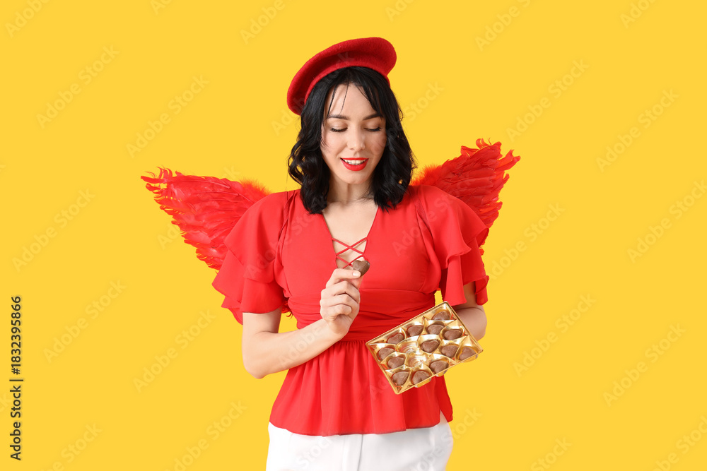 Fototapeta premium Young happy woman dressed as cupid with heart-shaped chocolate candies on yellow background. Valentine's Day celebration