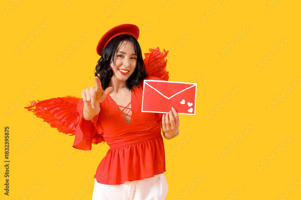 Obraz premium Beautiful young happy woman dressed as cupid with love letter pointing at viewer on yellow background. Valentine's Day celebration