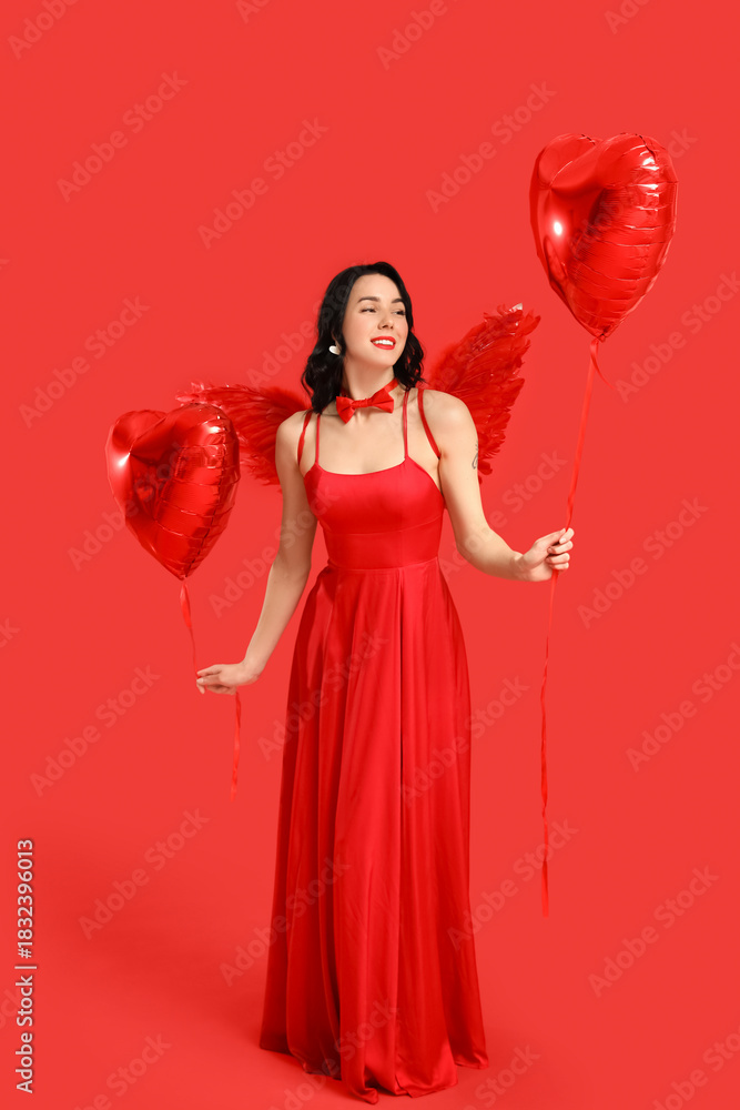 Fototapeta premium Beautiful young happy woman dressed as cupid with heart-shaped balloons on red background. Valentine's Day celebration
