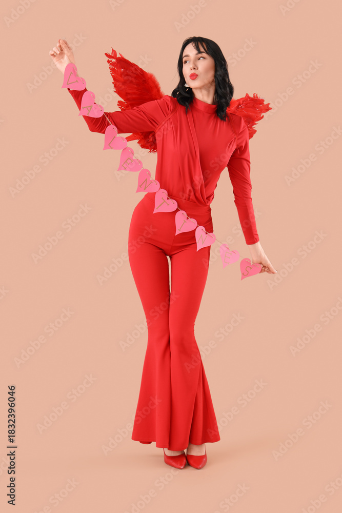 Obraz premium Beautiful young happy woman dressed as cupid with heart-shaped balloons blowing kiss on beige background. Valentine's Day celebration
