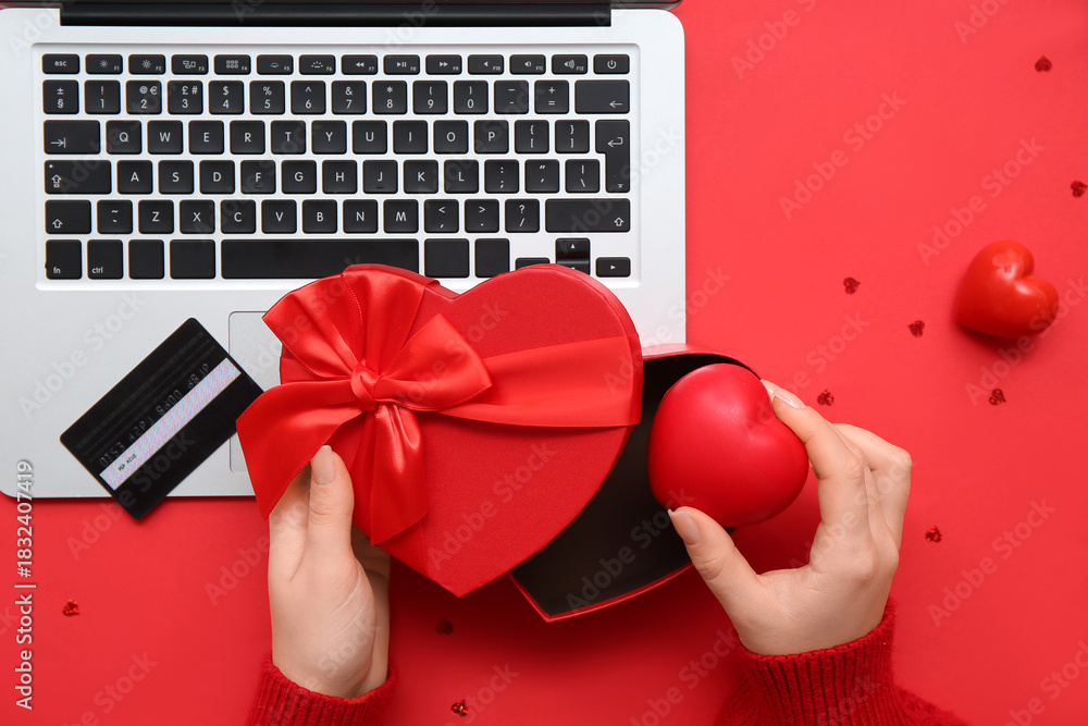 Fototapeta premium Female hands with gift box, heart, credit card and modern laptop on red background, closeup. Valentine's Day celebration