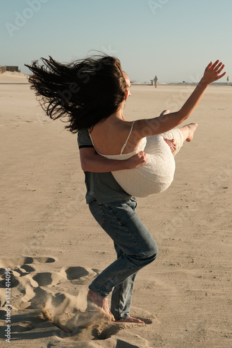 Man carrying woman in his arms on sandy beach, her hair flowing in the wind during a romantic and playful moment.