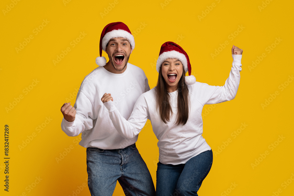 Naklejka premium Two excited individuals wearing Santa hats celebrate the Christmas season. They are full of joy and enthusiasm, standing together in front of a vibrant yellow backdrop.