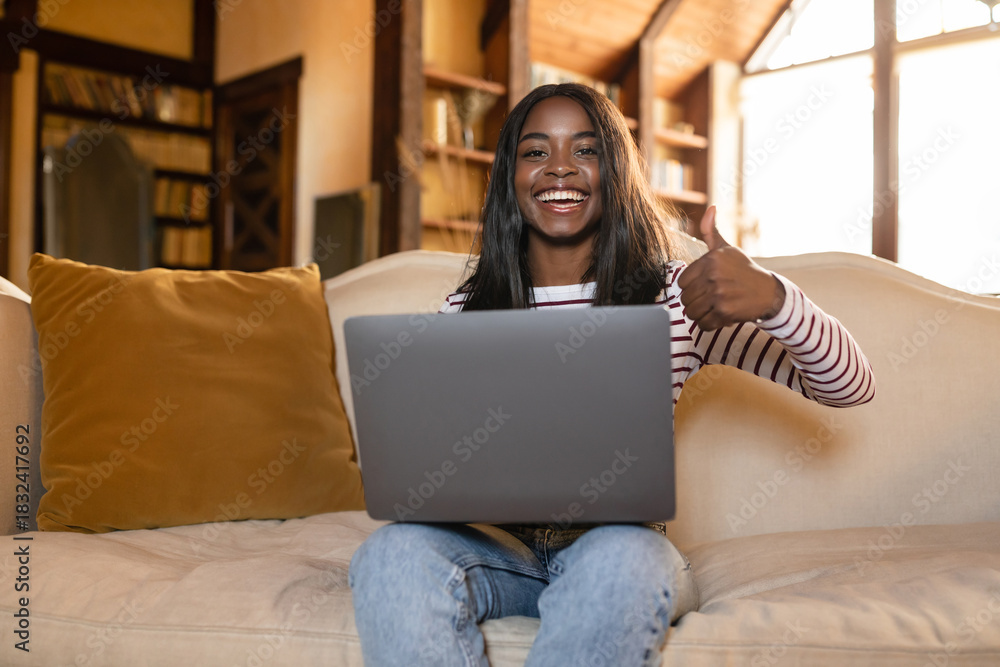 Fototapeta premium A woman sits comfortably on a couch in a bright living room, smiling and giving a thumbs up while using her laptop. Sunlight pours in through large windows.