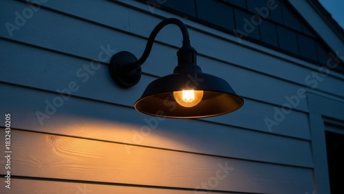 An old-fashioned outdoor wall-mounted light fixture with a warm incandescent bulb illuminates a white-sided house at dusk