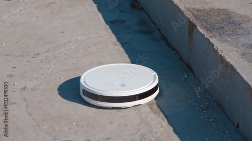 A round white robotic vacuum cleaner rides on a concrete surface outdoors at the edge of a raised platform, with shadows and small patches of grass visible nearby. uprising machines
