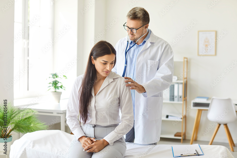Fototapeta premium Serious doctor checking up female patient in clinic holding stethoscope during medical exam, male therapist practitioner helping examines lungs or heart sounds, healthcare and health insurance
