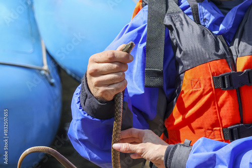 Close up of focused person hand holding rope, wearing blue jacket and orange life vest. This adventurer prepares for thrilling river rafting trip on sunny day